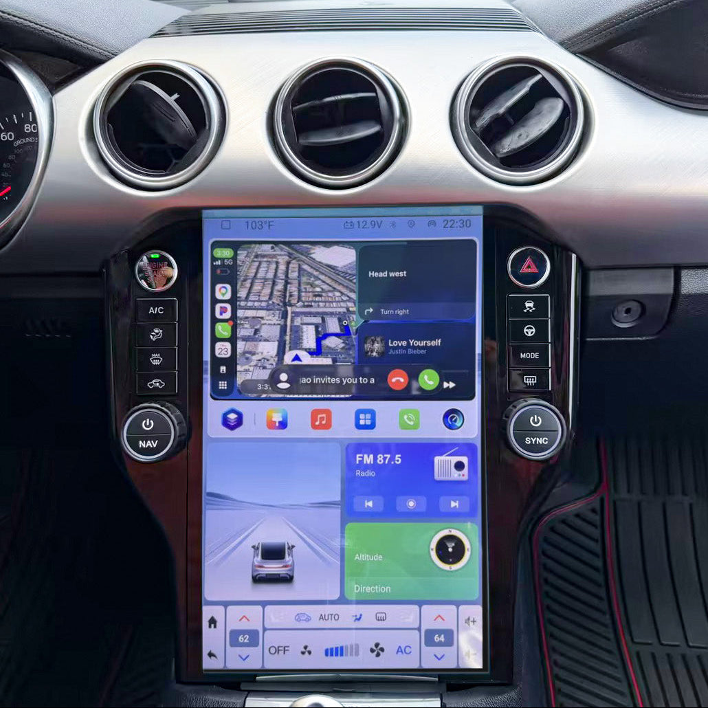 A large vertical touchscreen infotainment display mounted in the center console of a car interior, showing navigation and media apps. The dashboard around it is black and silver, with circular air vents and control buttons on either side of the screen.