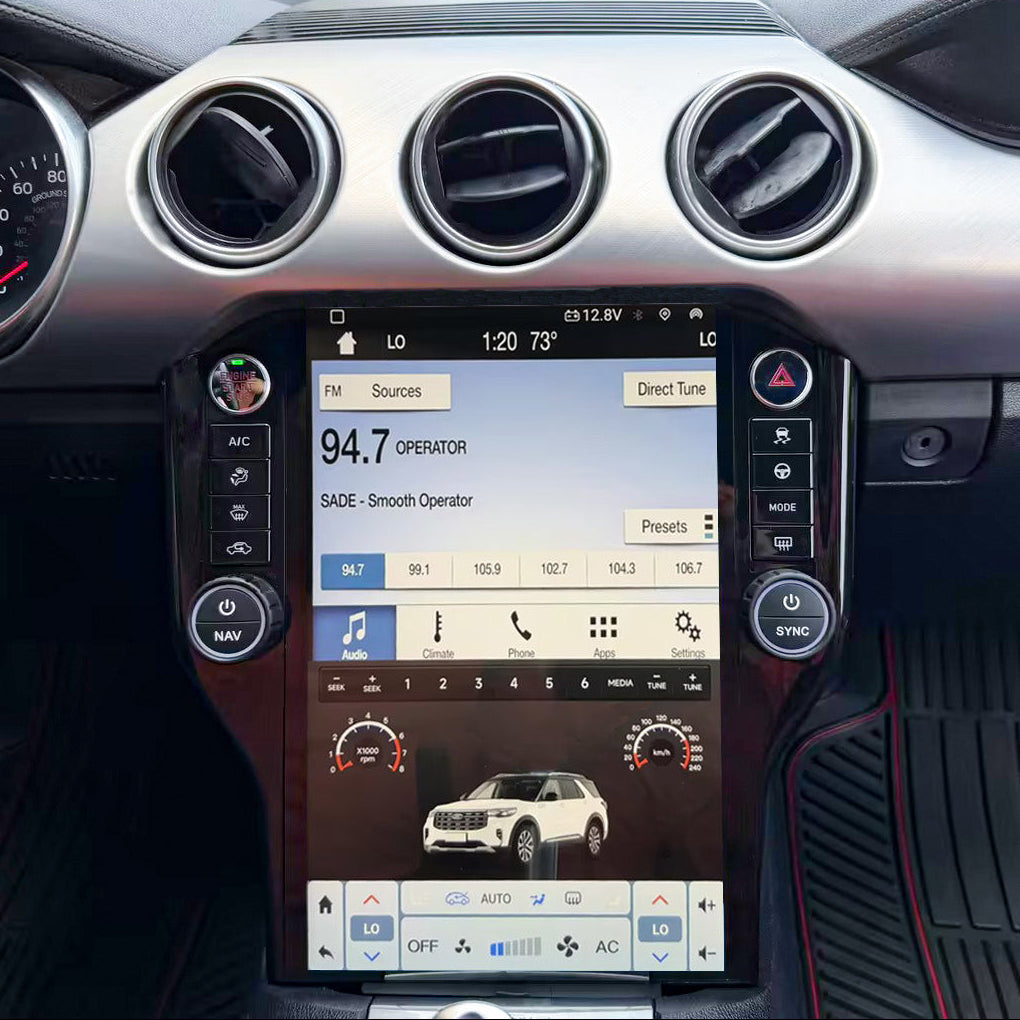 A large touchscreen infotainment display mounted in the center console of a car interior, showing a vehicle control interface with climate, audio, and vehicle graphics. The dashboard, air vents, steering wheel, and floor mats are visible around it.
