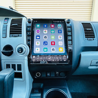 A large touchscreen infotainment system mounted on the center console of a vehicle interior, showing a grid of app icons on the screen. The dashboard, steering wheel, gear shift lever, and center console are visible in gray and black tones.
