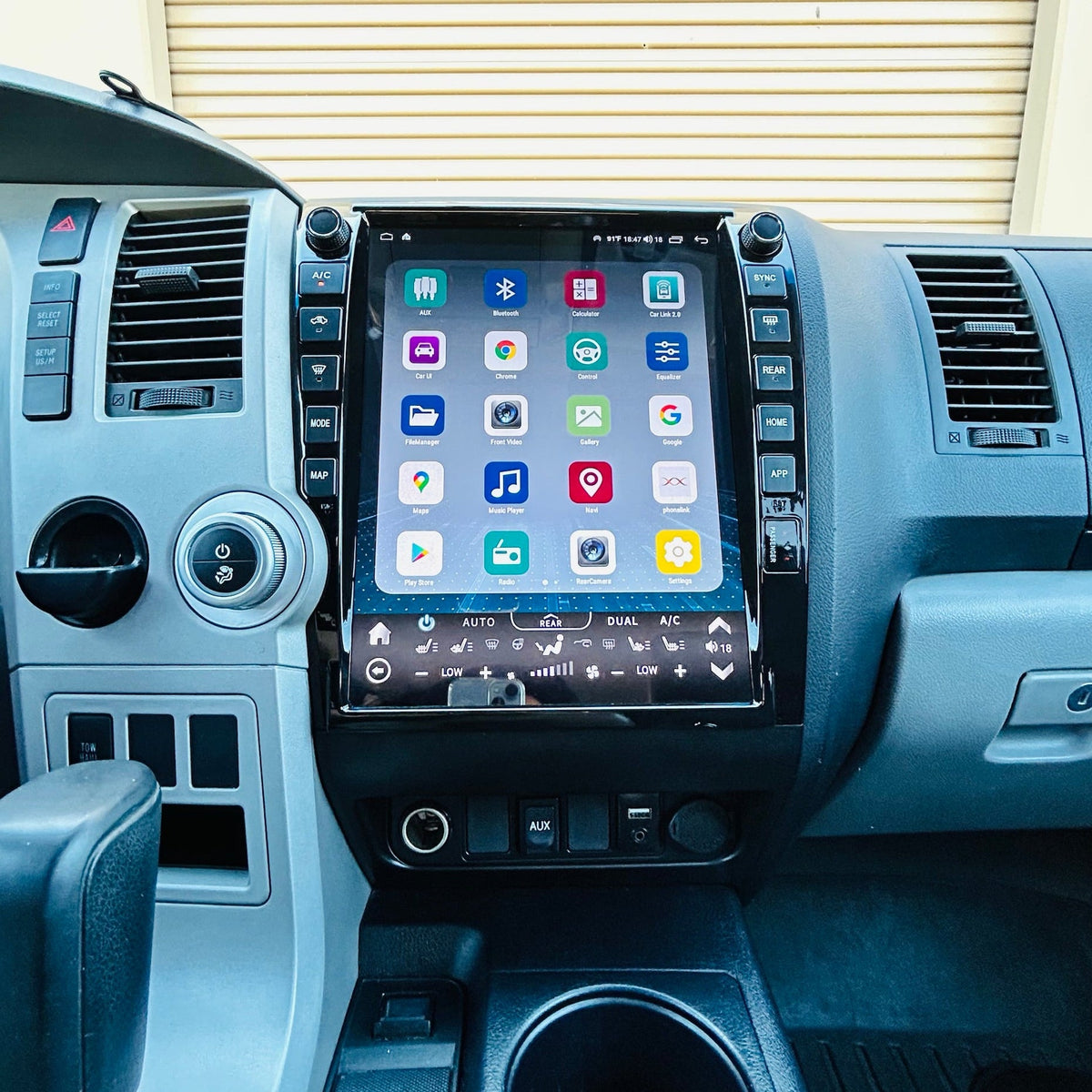 A large touchscreen infotainment system mounted on the center console of a vehicle interior, showing a grid of app icons on the screen. The dashboard, steering wheel, gear shift lever, and center console are visible in gray and black tones.