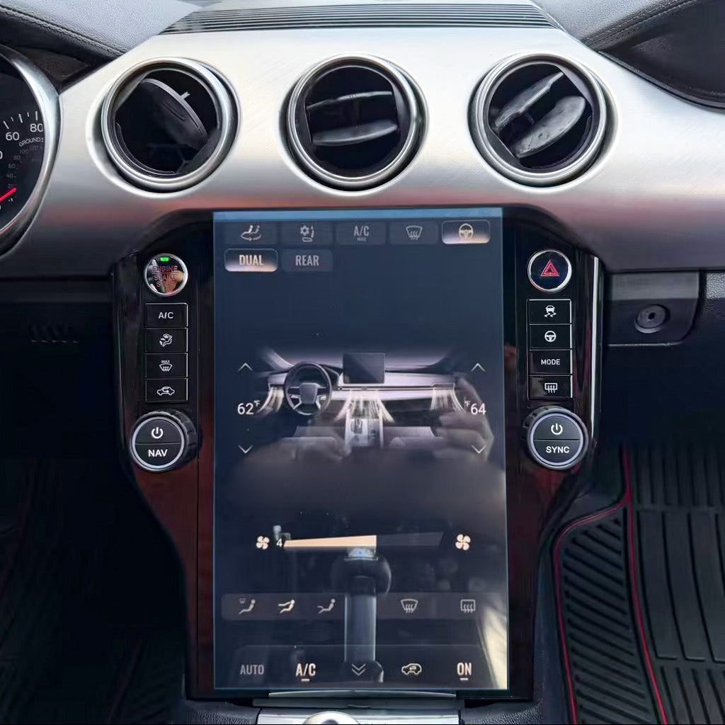 A close-up of a car's dashboard interior featuring a large vertical touchscreen display in the center console, circular air vents, silver trim, and black footwells with red stitching.