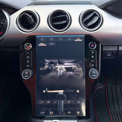 A close-up of a car's dashboard interior featuring a large vertical touchscreen display in the center console, circular air vents, silver trim, and black footwells with red stitching.