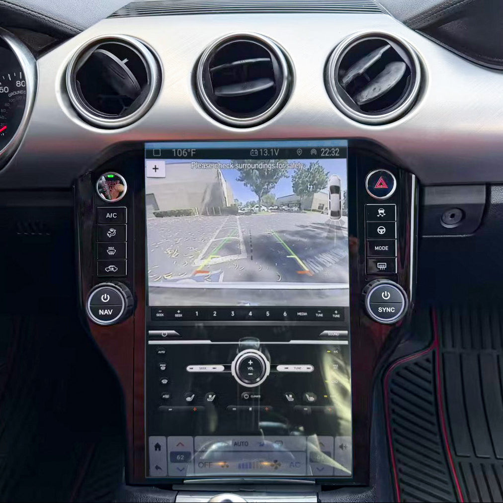 A close-up view of a car's dashboard interior, centered on a large vertical touchscreen display showing a rear-view parking camera feed. The dashboard has a silver metallic finish with three circular air vents, black trim, and dark textured floor mats with red stitching.